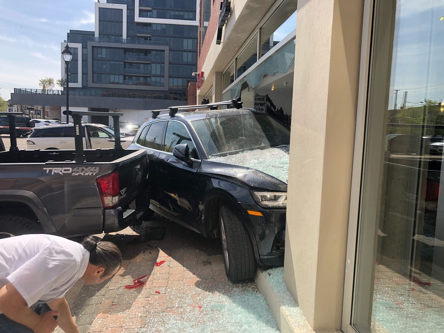 Photograph of an Audi SUV that has crashed through the front window of the Faema showroom. In the foreground, a man is bent over as if picking something up. There is shattered glass everywhere. 