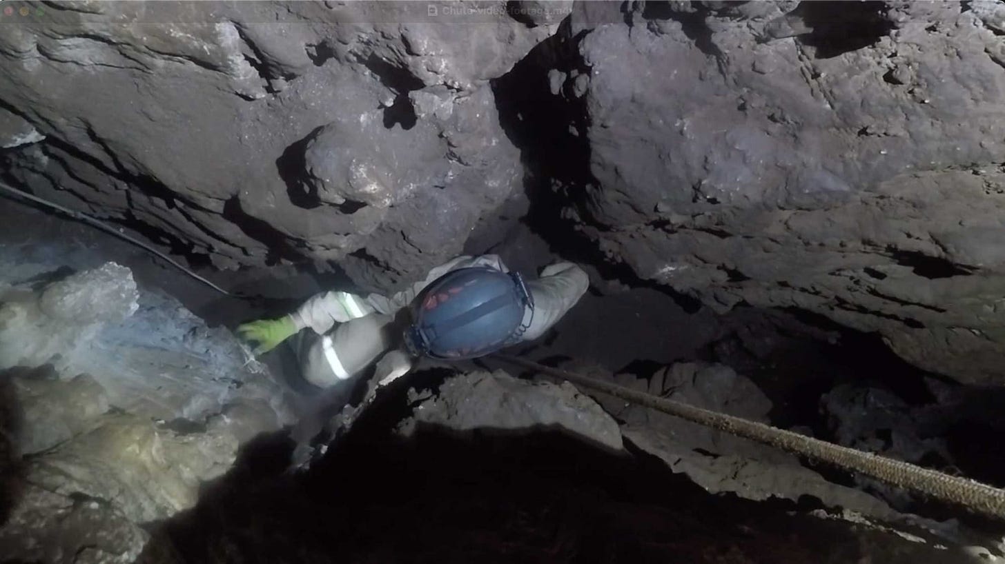 Helmeted caving person visible down within a very narrow fissure with ropes