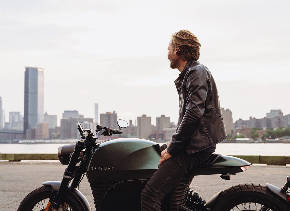 A Tarform Luna electric motorcycle and its rider with a city background. 