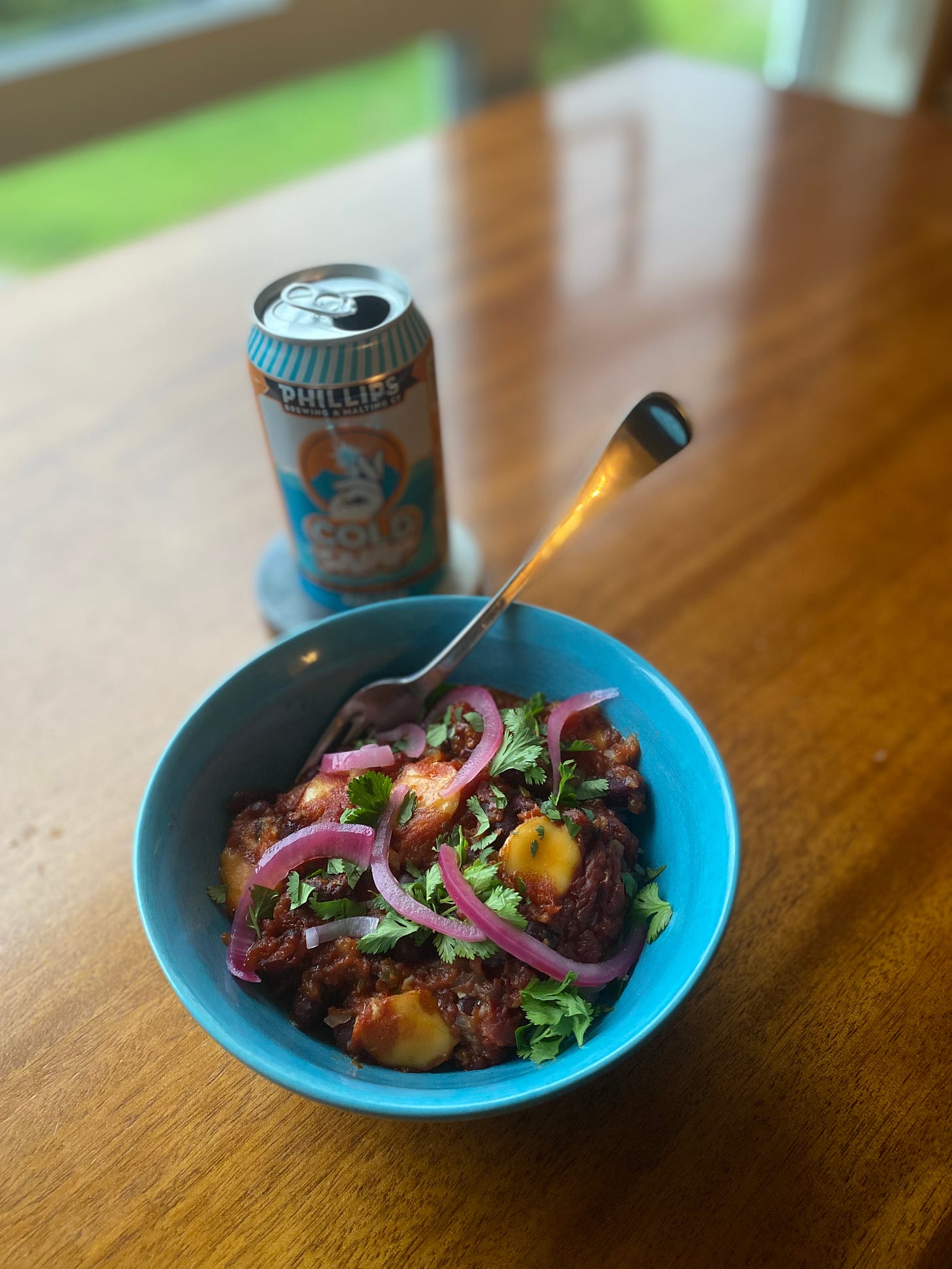 A blue bowl of the curry described above, deep red with pieces of mozza. Cilantro and pickled onion slices cover the top, and a fork sticks out at angle at the back. On a coaster behind it is a can of Phillips Cold Snap kolsch.