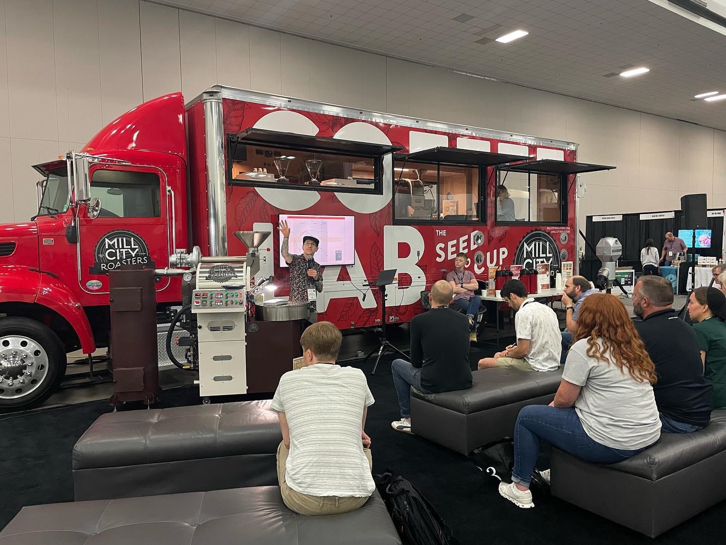 A man holding a mic gestures towards the ceiling while standing next to a coffee roaster in front of a bright red box truck semi that has windows cut into the side to expose the coffee brewing machines inside. An audience sitting on grey cushioned benches takes notes with their back to the camera.