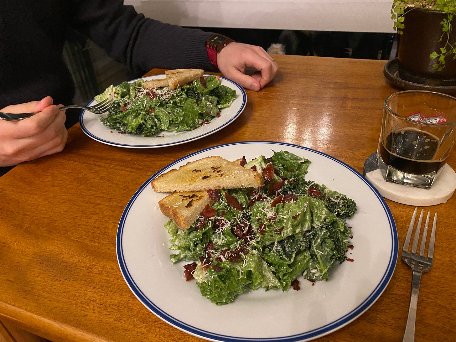 Two white plates with blue rims, each with two halves of diagonally sliced garlic toast laid over caesar salad sprinkled with parmesan and crumbled salami. On a coaster is a mostly-empty glass of porter. Jeff's hands are on the table next to his plate, one holding his fork just above it.