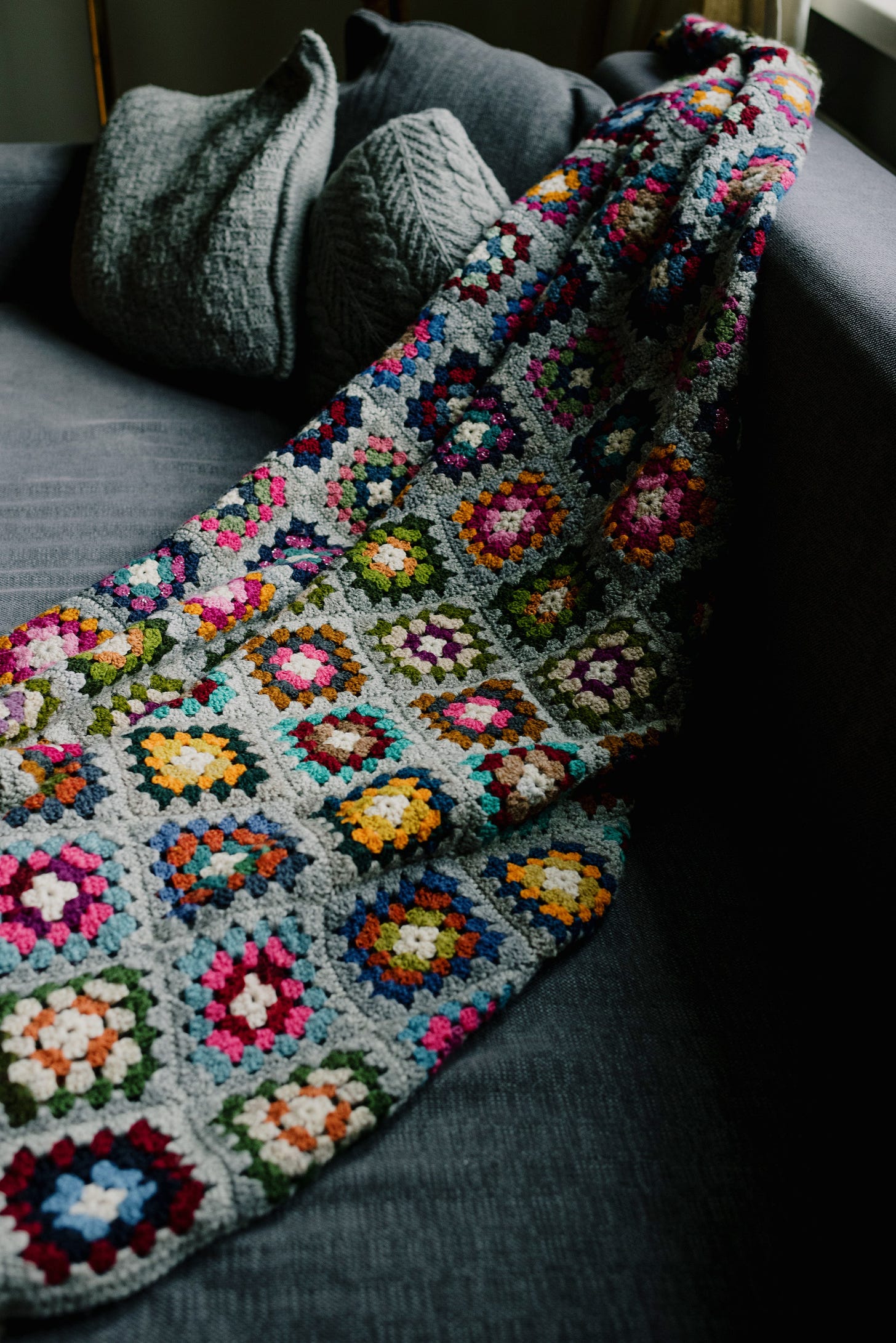 A colourful crocheted blanket lies across the cushion and over the back of a grey sofa.