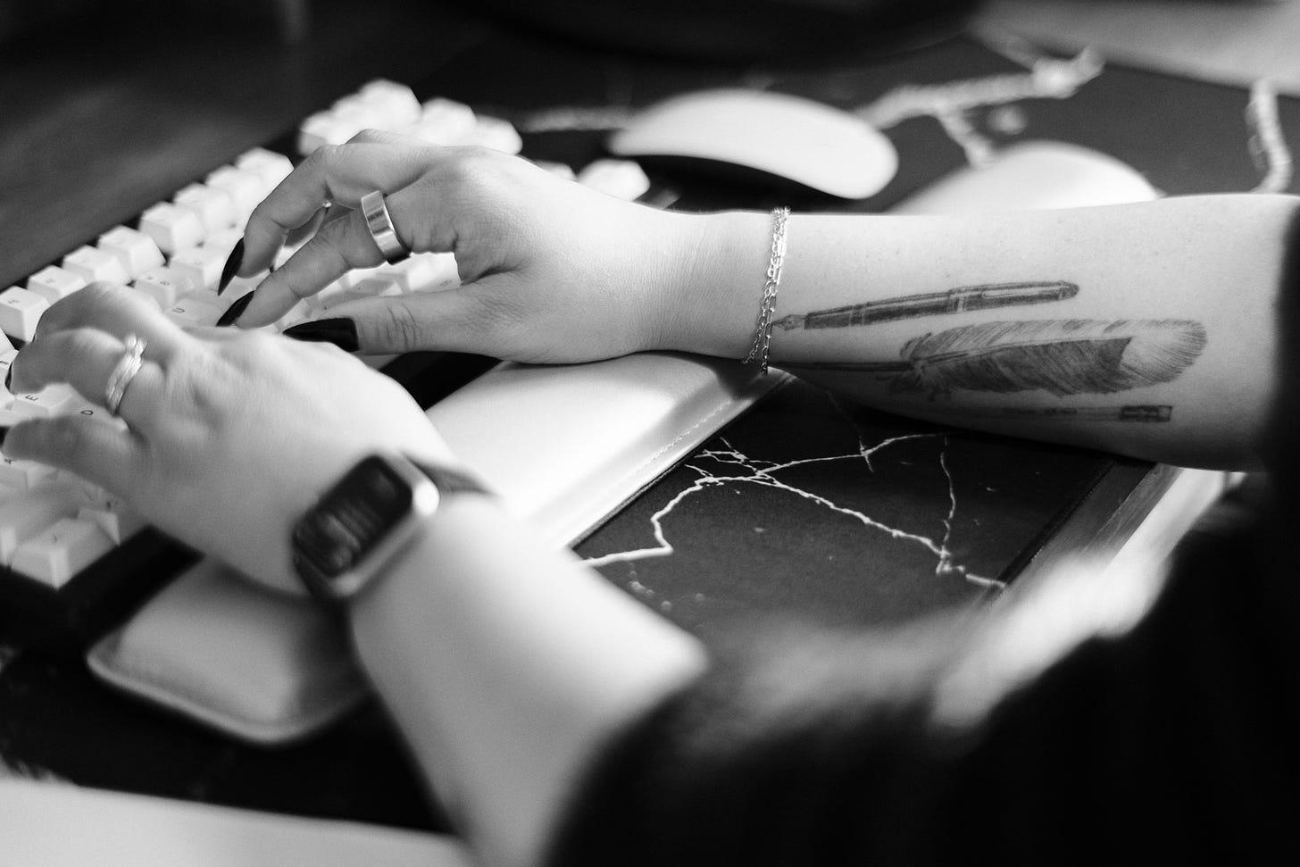 black and white photo of a woman's forearms as she types on a keyboard. she has long black nails and wears rings, bracelets, and an Apple Watch. she has a tattoo on her right forearm of a pen, feather quill, and pencil.
