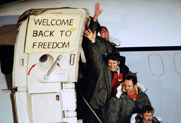 Several Americans in olive parkas exit an airplane, some of them smiling and waving. A sign that reads “Welcome back to freedom” is attached to the door.