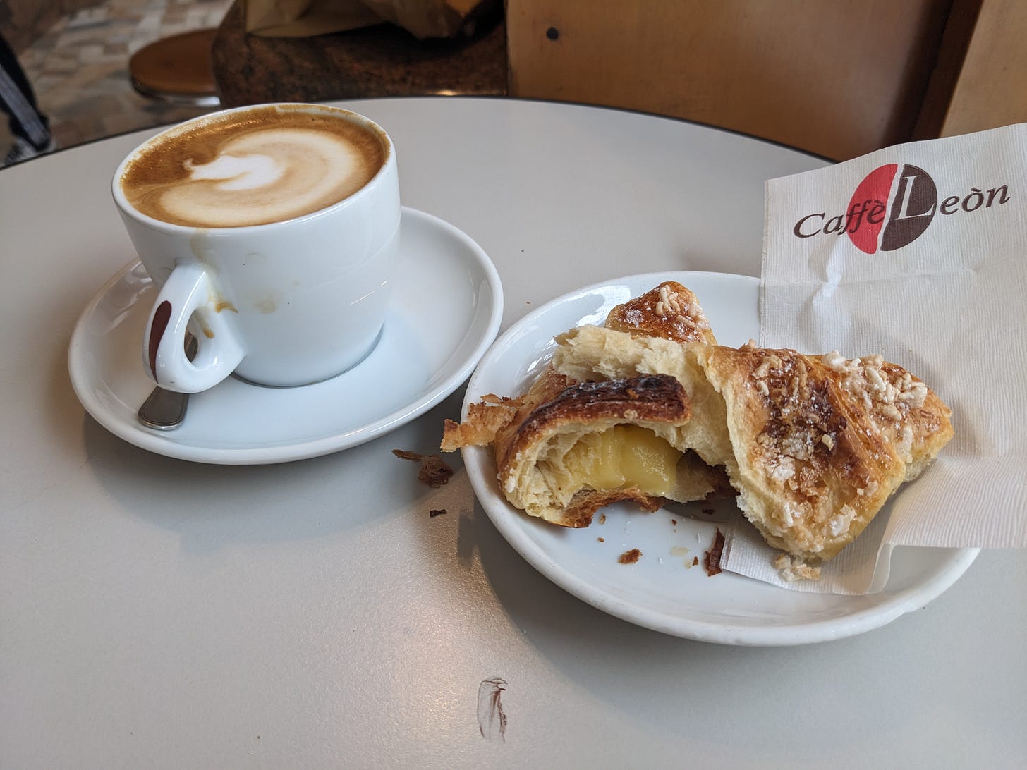 On a table sits a cup of coffee in a white cup, and a pastry which has been torn in half