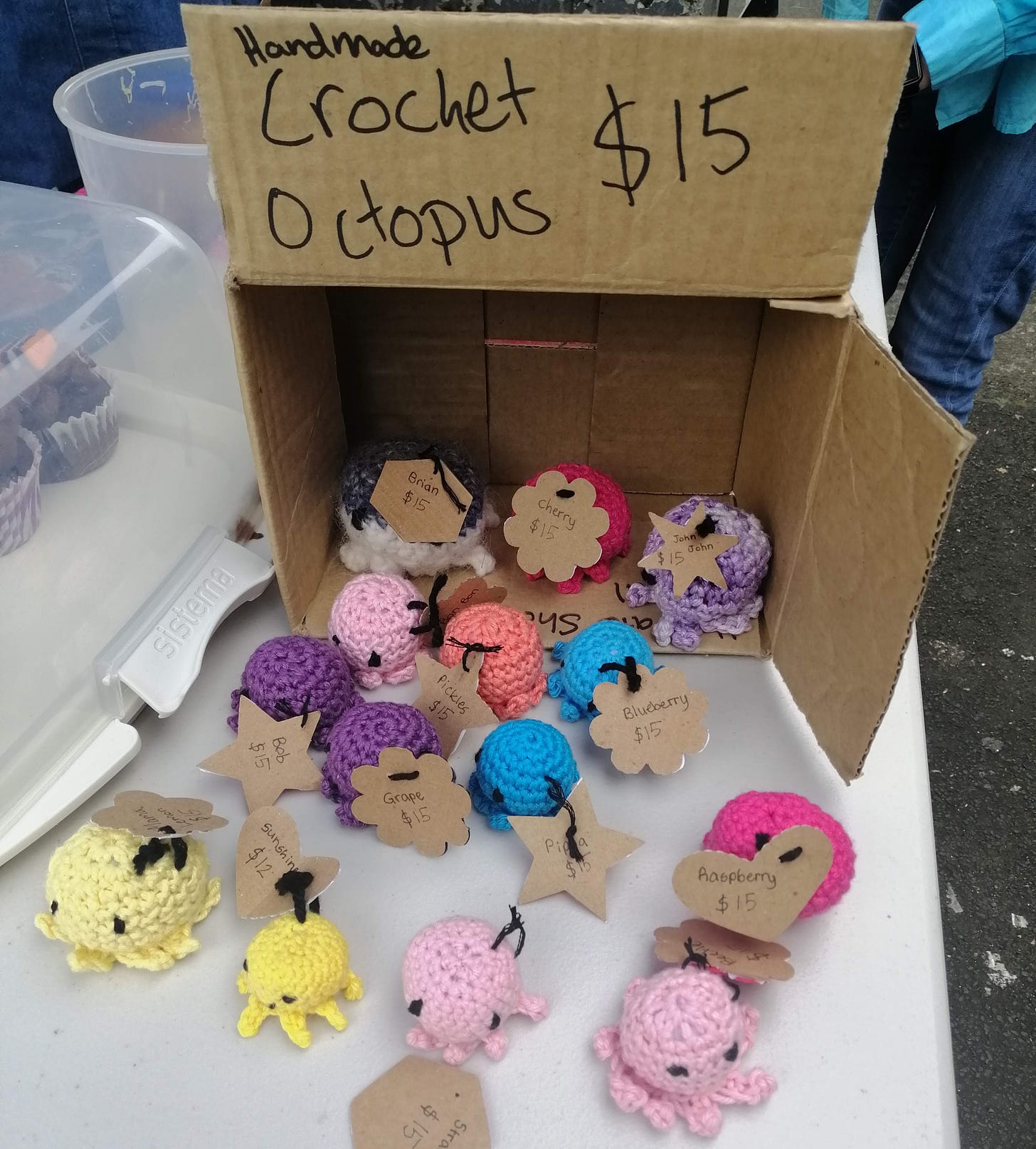 A box with a sign that says "handmade crochet octopus $15" and a consortium of crochet octopi on the table beside cupcakes for sale. 