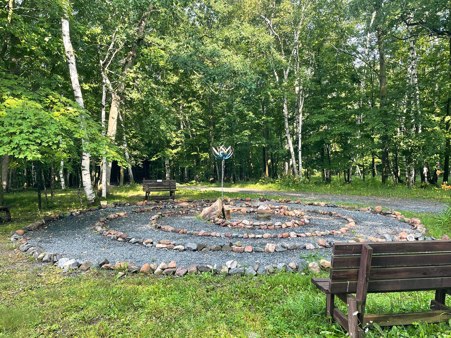 Birch trees in background behind a circular labyrinth outlined in rocks and bordered by two brown benches.