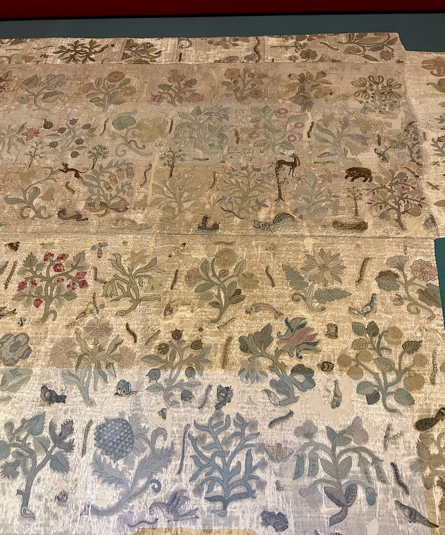 A grey looking piece of fabric embroidered with plants, and small scale animals including a bear, a deer, and a hunting dog, alongside insects and caterpillars