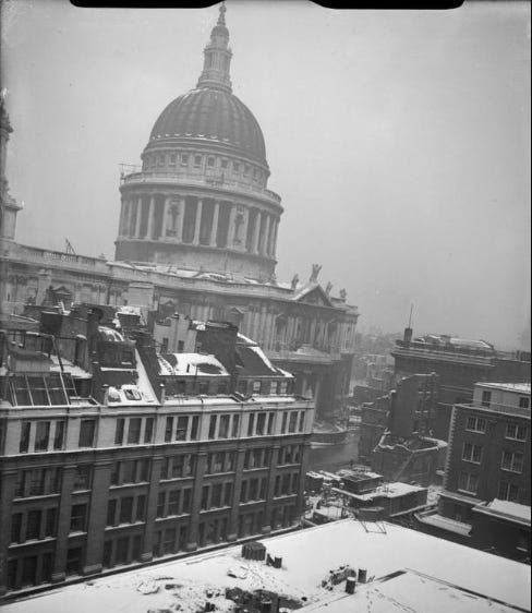 Black and white photo of the dome of St Paul's Cathedral taken from the roof of a nearby building: snow is on the ground and roofs, and there is a hole and broken walls where a building once stood