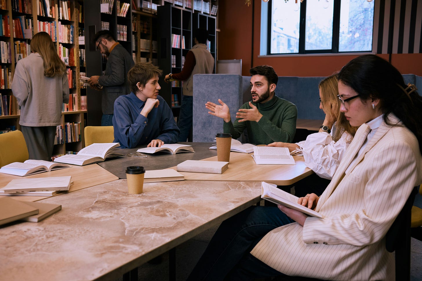 A group of writers sitting a table in a library. There are books on the table and coffees, with people browsing books in the background. 