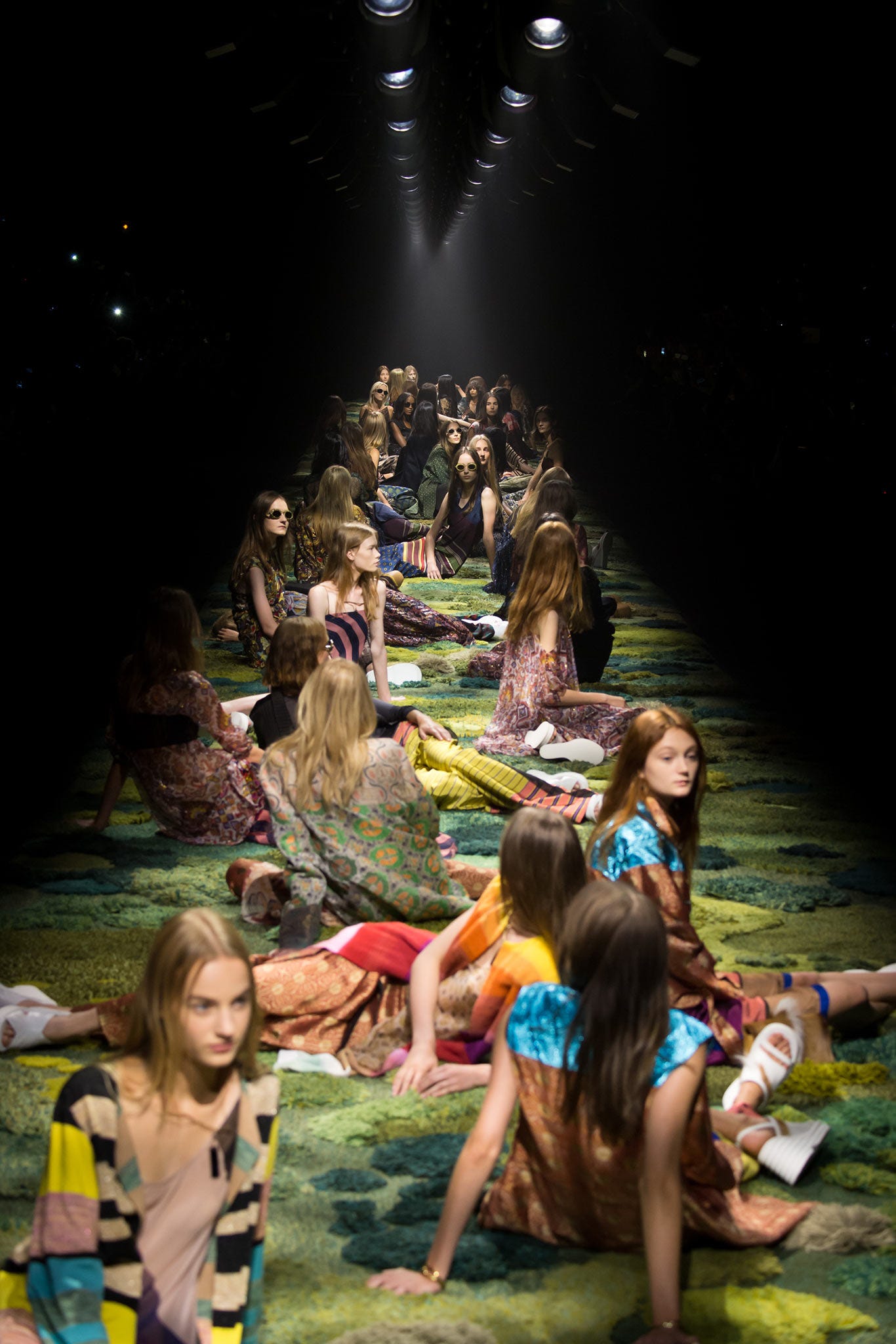A dimly lit catwalk with spotlights that focus on over 30 women models, who are all elegantly dressed and seated on a thick textured green carpet that is deliberately made to resemble grass. A dimly lit catwalk with spotlights that focus on over 30 women models, who are all elegantly dressed and seated on a thick textured green carpet that is deliberately made to resemble grass.