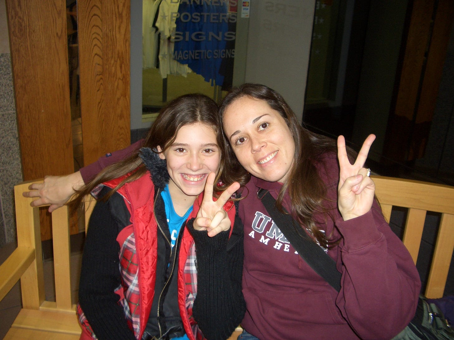 Two people sitting on a bench showing the Peace sign with thier hands. One is wearing a burgundy sweatshirt with UMASS Amherst written on the front. 