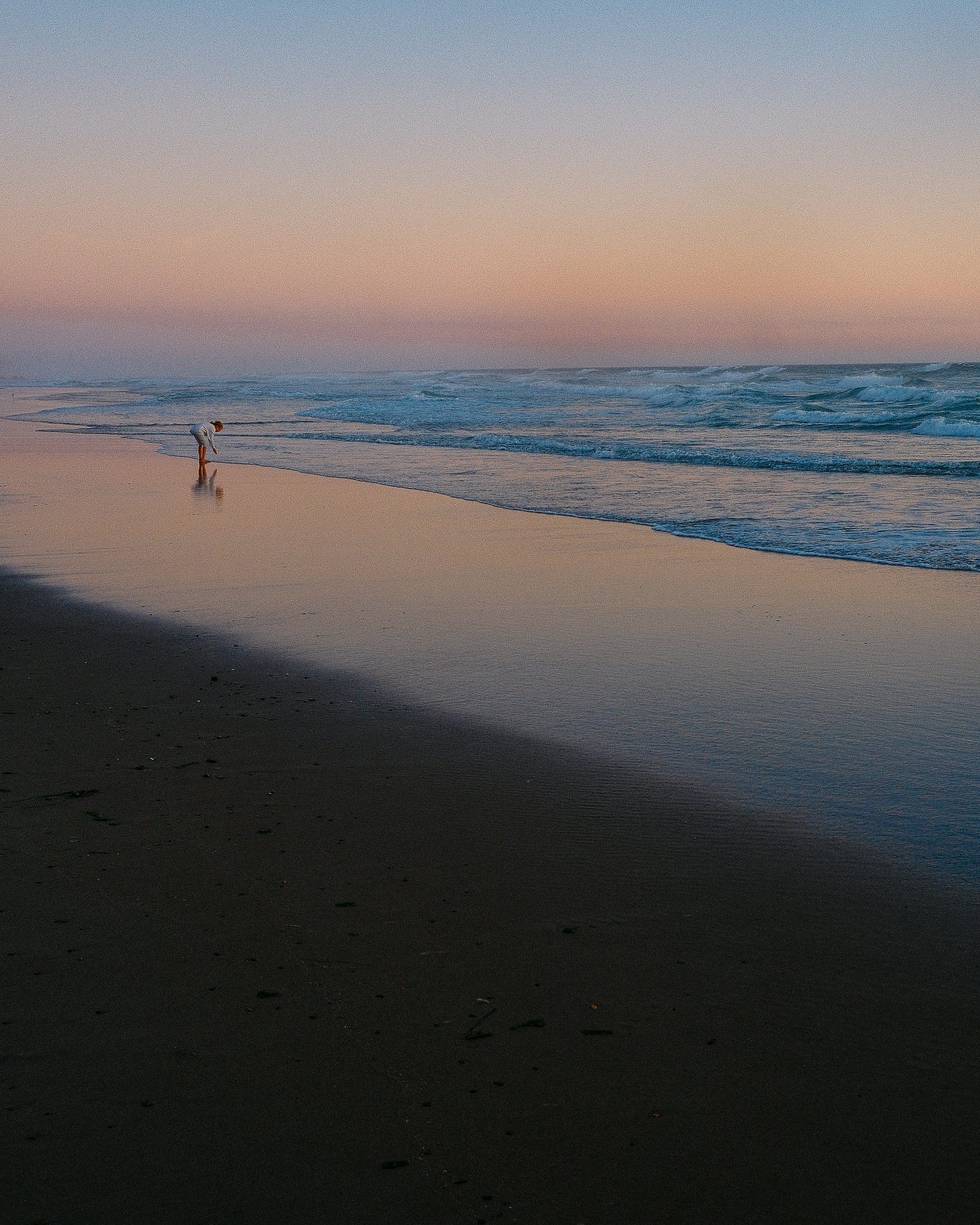 a person stands on the ocean shore, with waves, and a pink/yellow color sunset reflecting off the water, and dark sand