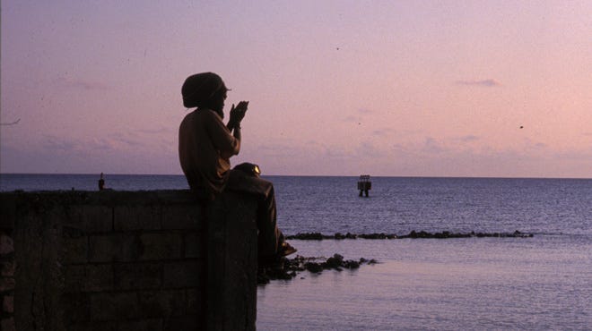 A sunset image of a Rastafarian man sitting on a jetty looking out at the sea.