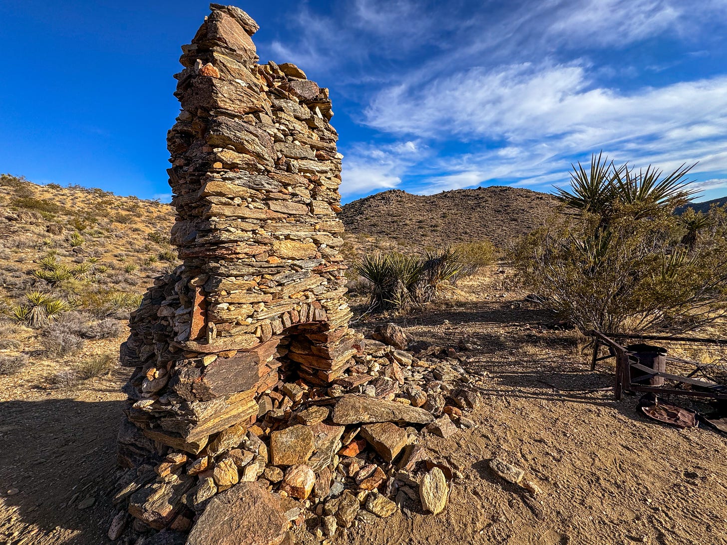 A stone fireplace rises from the desert floor without any structure around it. A Mojave yucca is directly behind it, and two small hills are further out.