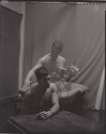 Photo en noir et blanc de deux hommes nus. Le premier est assis à table. Le second est derrière lui, son corps est transparent et il tient dans une main des fleurs et s'apprête à toucher l'épaule de son compagnon de l'autre.