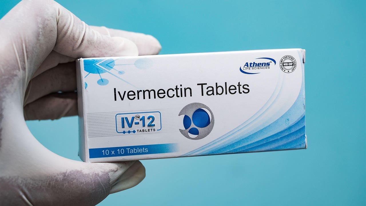 A white rubber gloved hand holds a box of ivermectin tablets