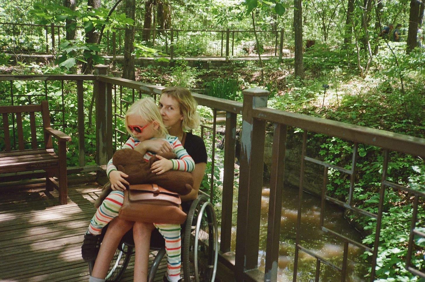 Rebekah sits in her wheelchair holding her son in her lap. She looks toward the camera, her nose pressed against the back of her son’s head. Wearing his favorite rainbow pajamas and red sunglasses, her son looks down and to his right. They are surrounded by trees and green foliage.