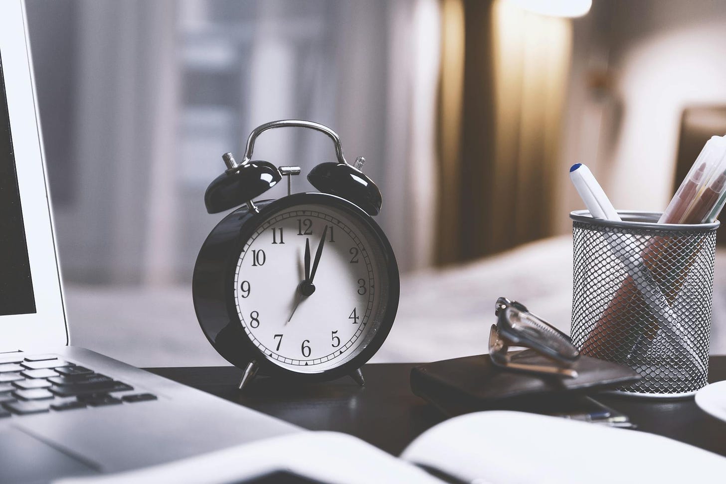 A clock on a desk in between the edge of a laptop and a pencil cup with pens in it