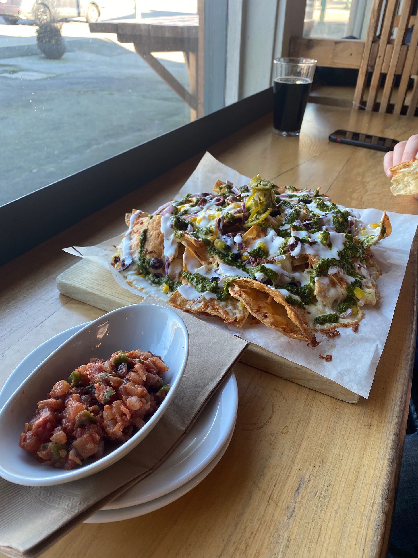 At a wooden bar near a window, the nachos described above, on parchment atop a wooden board: crispy chips drizzled with crema and chimichurri and covered with white cheese, slices of olives and jalapeño, and red onion. In a boat-shaped dish in the foreground is a dish of pico de gallo.