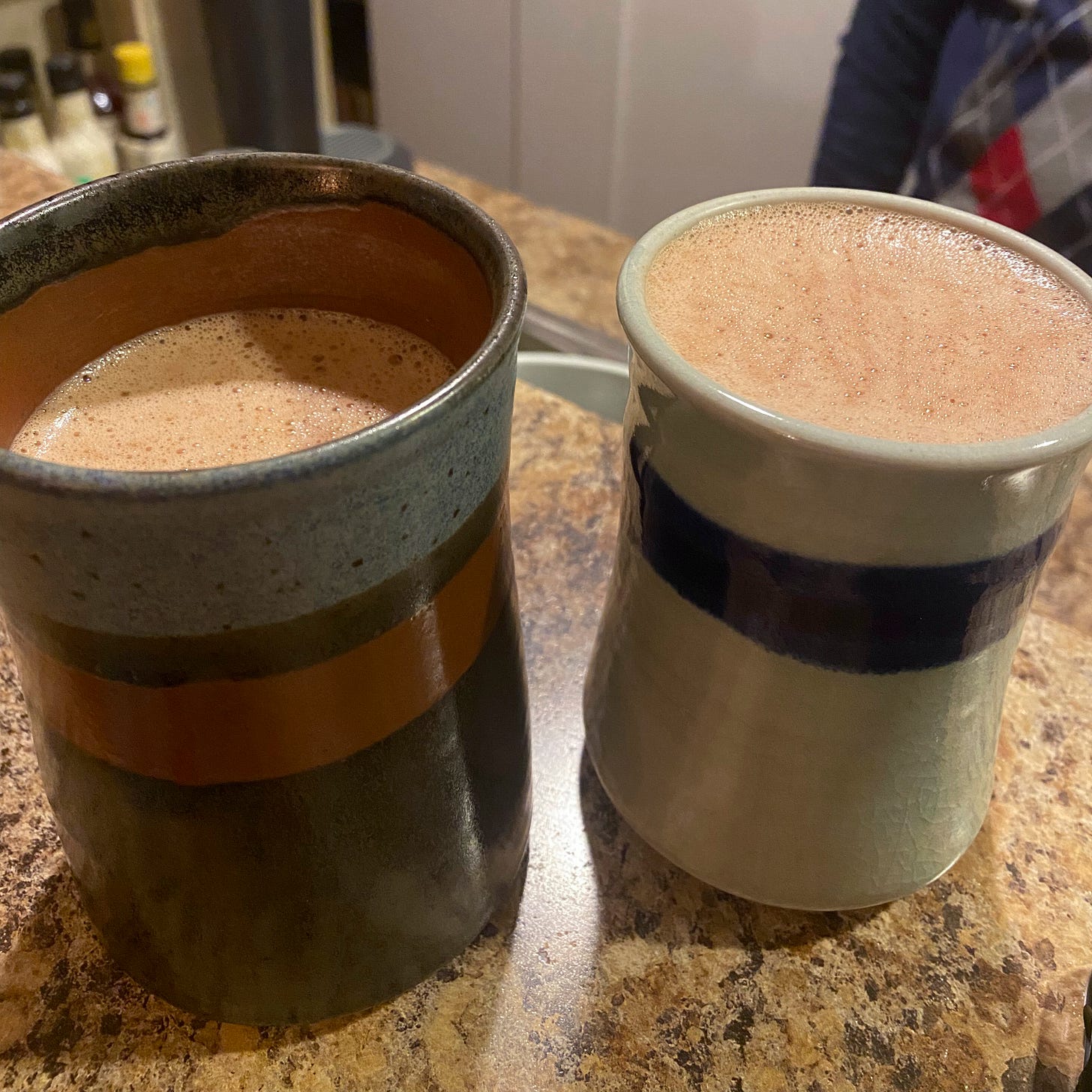 Two ceramic pottery mugs, mismatched but both with a painted stripe around it, filled with hot chocolate. The mug on the right is full to the brim, as it is a little smaller than the mug on the left.