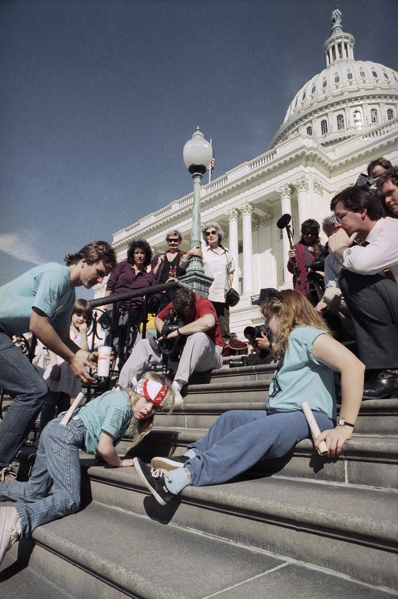 US Capitol building staicase, a young girl crawling up the stairs on her belly, surrounded by supporters and journalists taking photos and filming her, watched over by some passerby ladies in sunglasses.