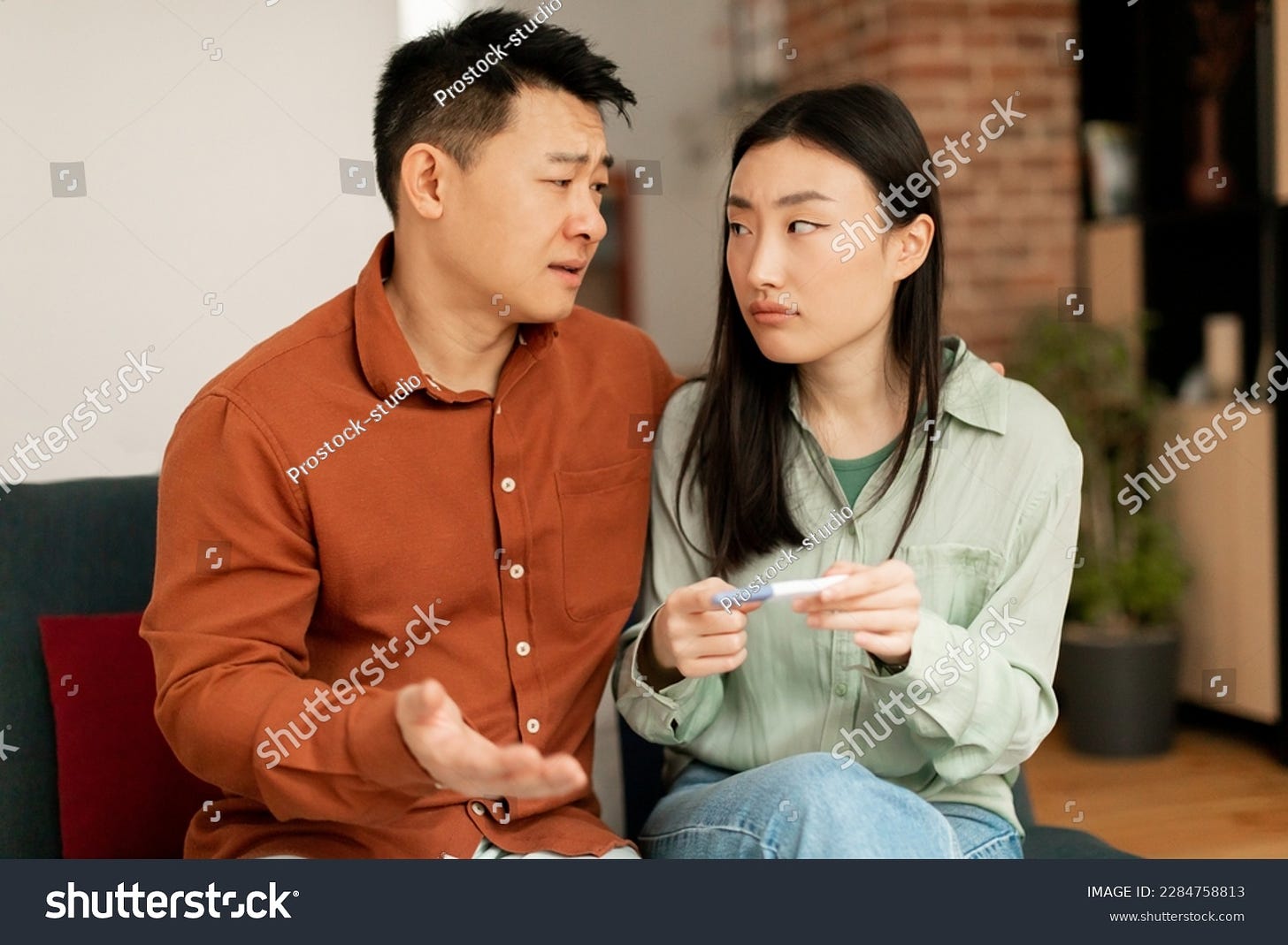 Depressed sad asian husband and wife upset of negative pregnancy test result, sitting on sofa in living room interior, copy space. Infertility, health problems and childlessness