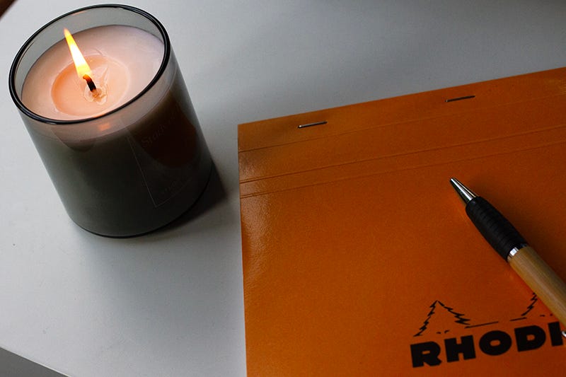 A Rhodia paper pad, pencil and a lit candle on a white desk.