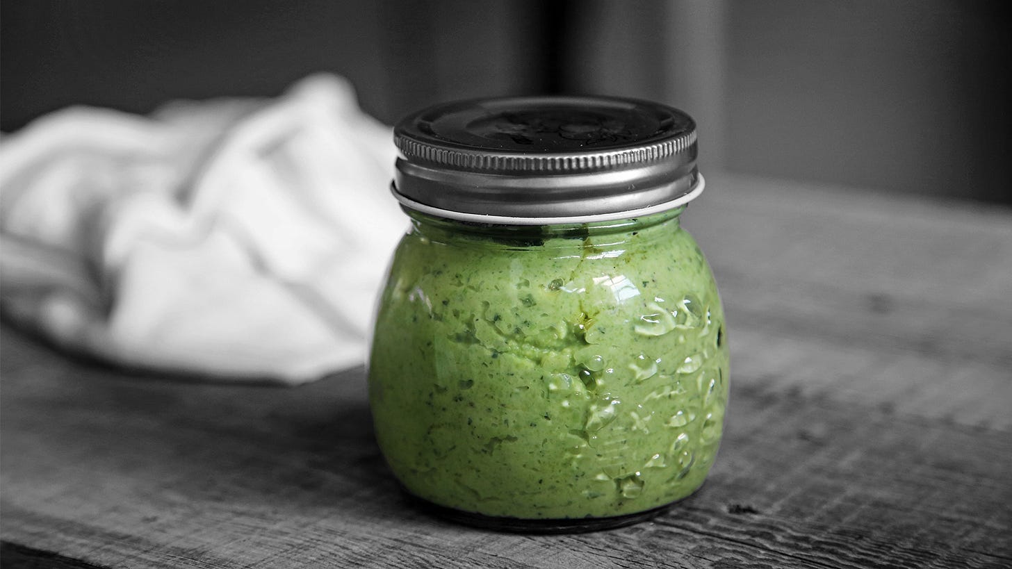 A small canning jar of pesto sauce stands proud on a rough wood surface