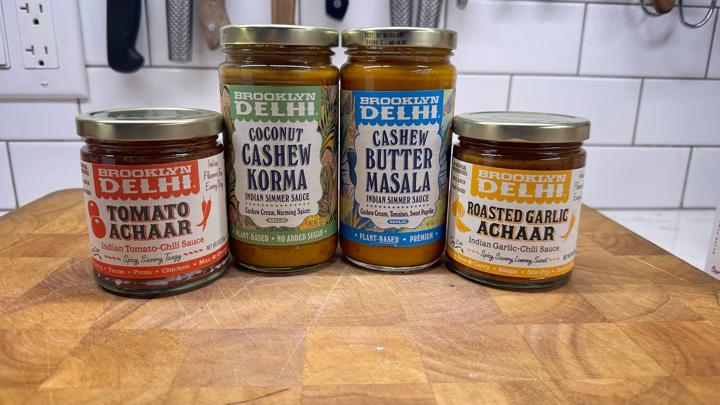 Four jars of Brooklyn Delhi products. From left to right: Tomato Achaar Indian Tomato-Chili Sauce, Coconut Cashew Korma India Simmer Sauce, Cashew Butter Masala Indian Simmer Sauce, and Roasted Garlic Achaar Indian Garlic-Chili Sauce