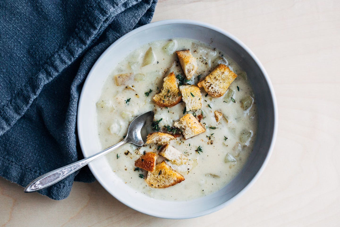 A bowl of potato soup with croutons and herbs. A spoon rests in the soup.
