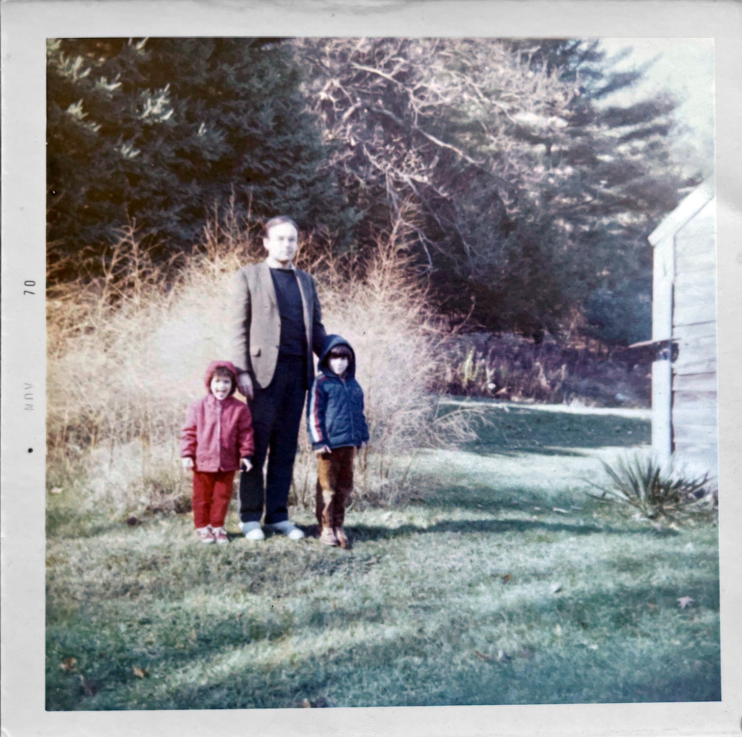 A man in a sport jacket stands flanked by two young children outdoors on grass by the side of a barn. 