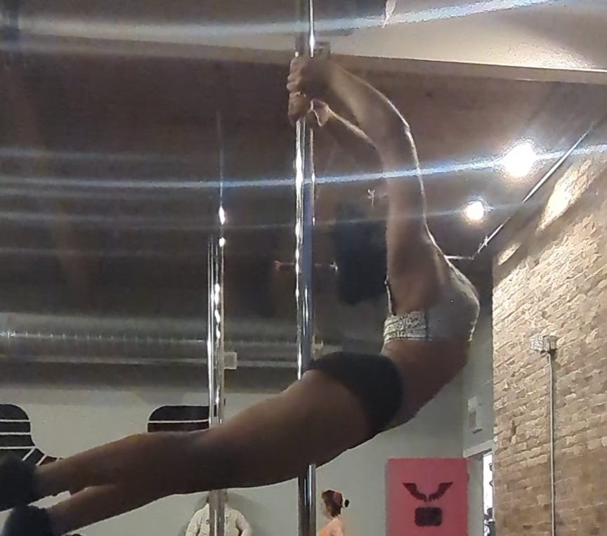 Nathalie is squeezing a metal pole between her legs as she wraps her hand above her head to grab the pole above her