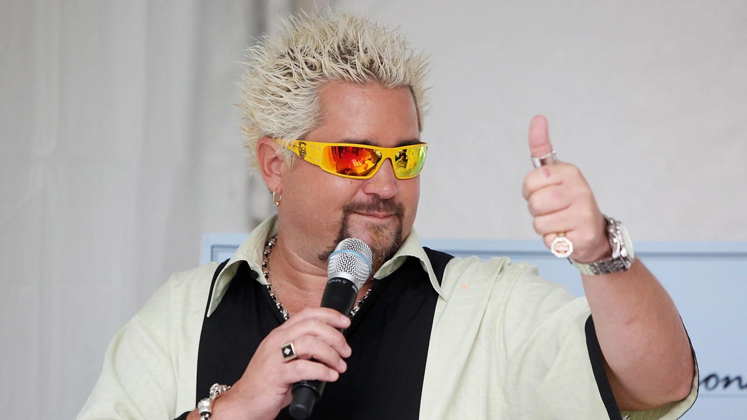 Guy Fieri in yellow sunglasses with orange reflective lenses giving a thumbs up. He's also holding a mic and wearing a bowling-type shirt with a big black stripe down the center; the arms and collar are cream colored.