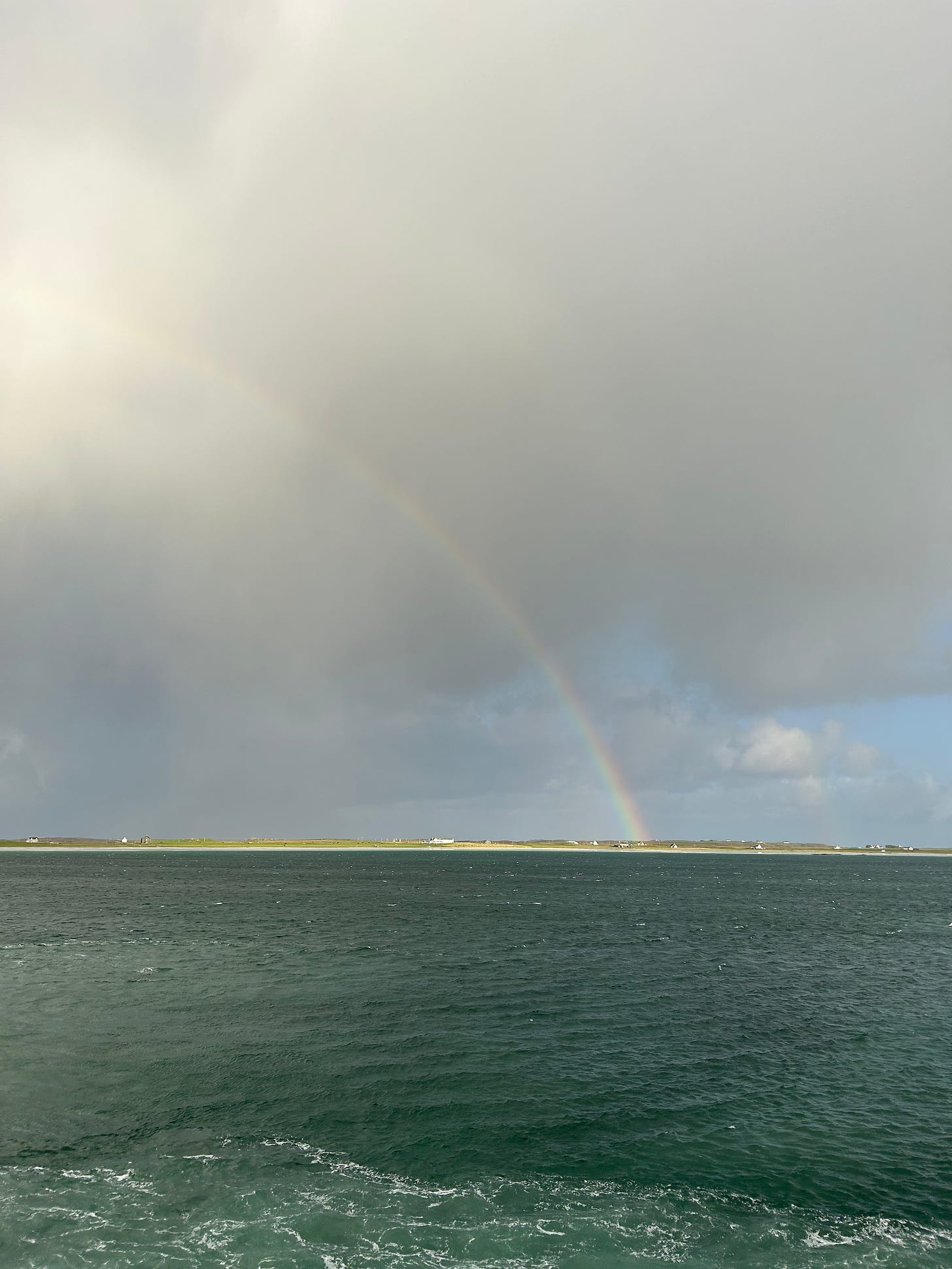 A rainbow arcs over a long, low green island dotted with white cottages. The sky around the rainbow is grey and stormy.