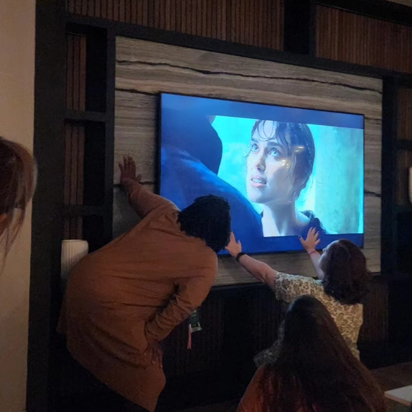 Julie Soto kneels in front of the TV, hands raised, as Nikki Payne looks on. The TV is showing Kiera Knightly's weird bangs as Darcy proposes in the rain.