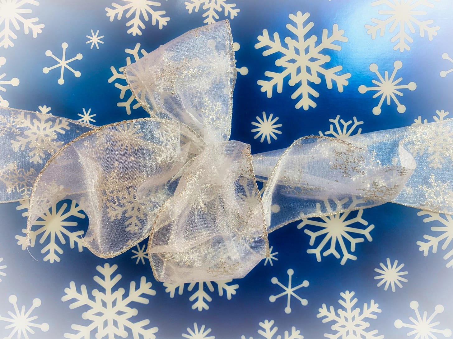 A close-up of a wrapped gift in blue metallic paper ewith snowflakes and a white sparkling ribbon tied into a bow