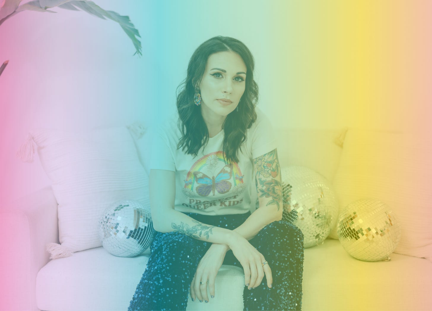 A rainbow-filtered photograph of Shohreh sitting on a couch, surrounded by glittering disco balls. She's wearing navy sequined pants, a tee with a butterfly and a rainbow on it, and she's staring directly into the camera with her arms crossed in her lap.