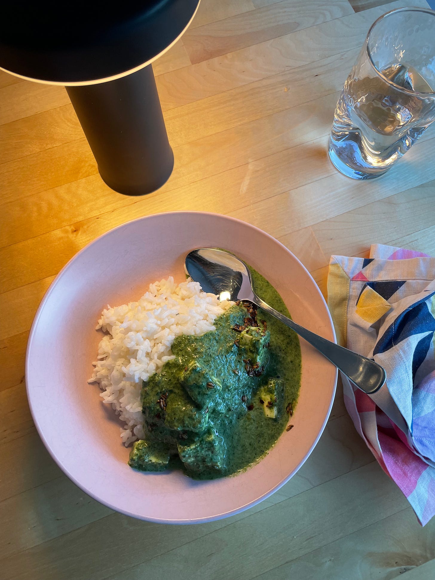 A pink bowl with rice and coconut spinach panner, and a wavy water glass to the side.