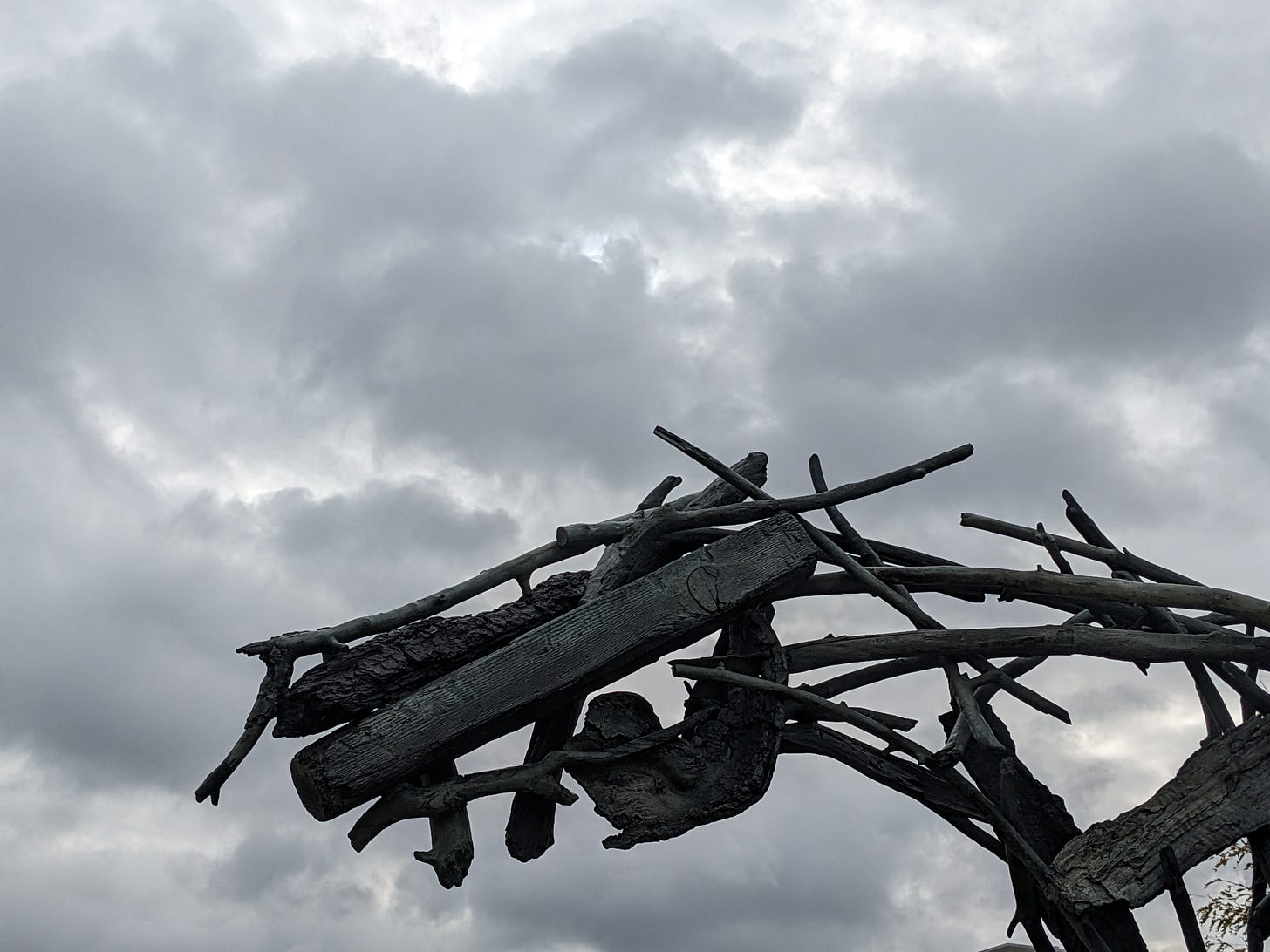 Photograph of a sculpture against a cloudy gray sky. The visible part of the sculpture is a horse head constructed from bronze twigs.