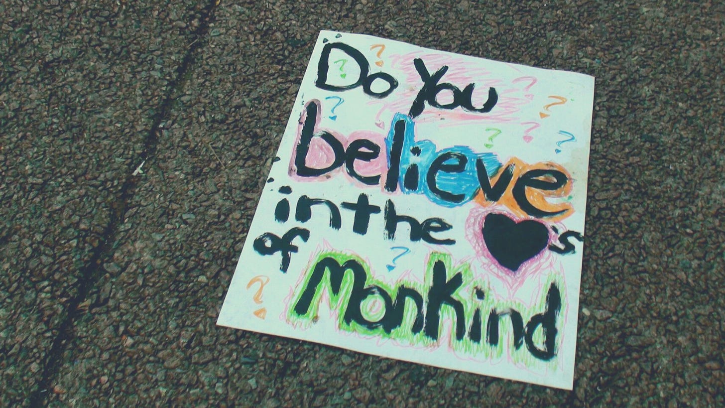 A protest sign on a cement and gravel ground. The sign is colorful and says in handwritten scrawl: "Do you believe in the hearts of mankind" with the word hearts as a drawn on heart.