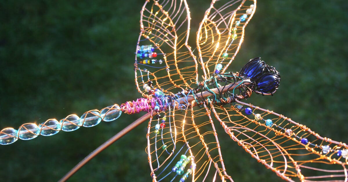 An image of a dragonfly made of copper wire and colorful beads pictured in front of a deep grassy green background. 