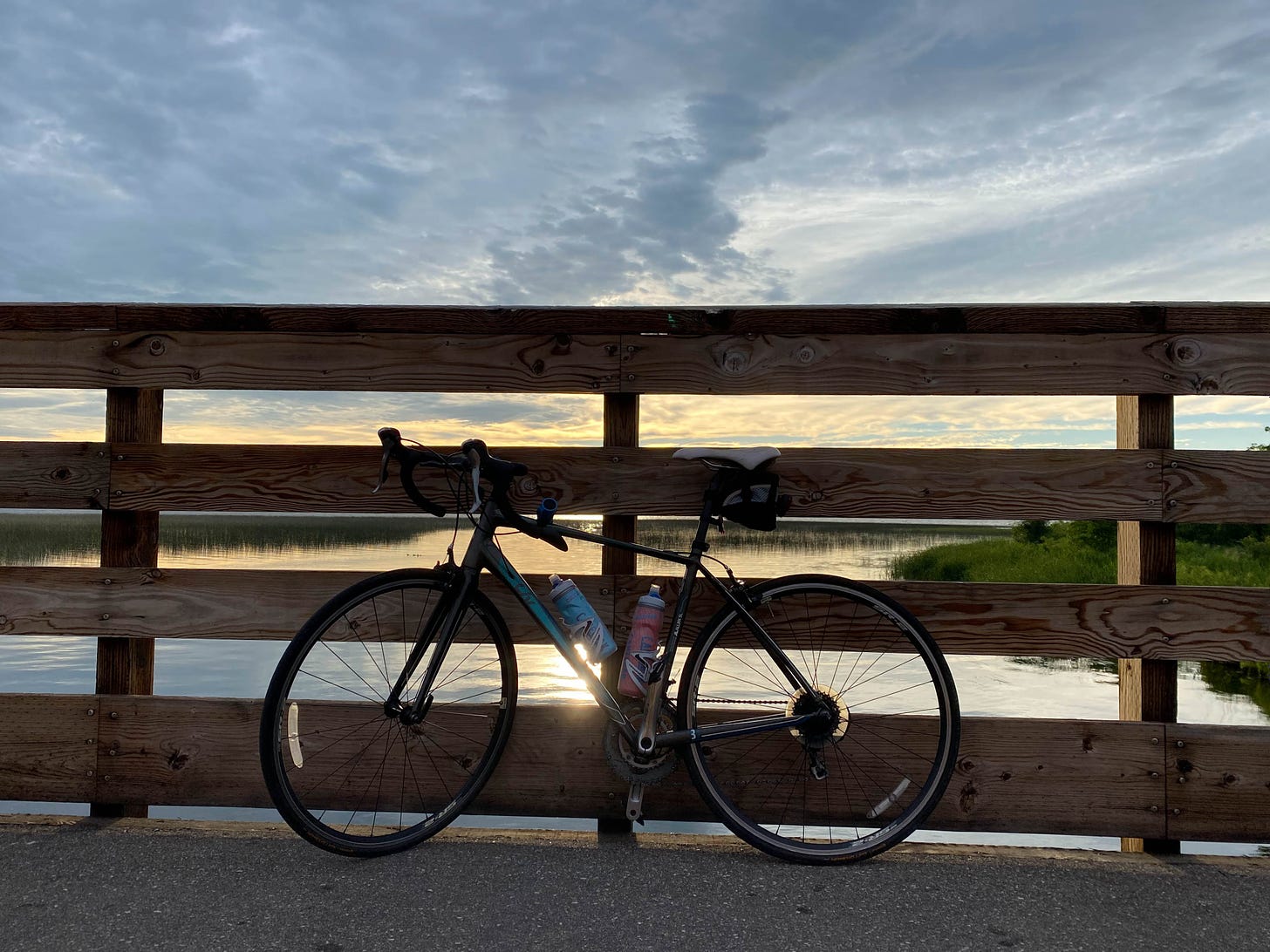 Silhouette of bike in front of wooden fence with lake, sun, and clouds in backgound.