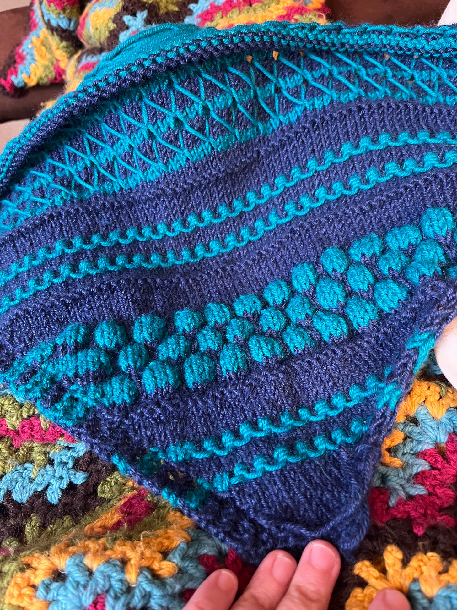 A hand holds down the corner of an item knitted on the bias - it has alternating bands of smooth stockinette stitch and highly-textured bubbles and elongated stitches. The background color is dark blue and the contrast color is teal, even though the obnoxiously bright blanket being used as a backdrop doesn't make it look that way.