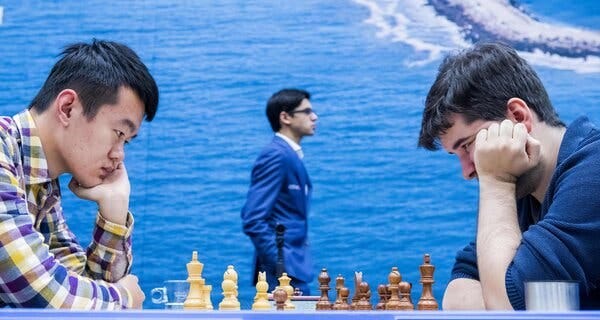 Ding Liren and Ian Nepomniachtchi sit across from each other, both with a hand on their face, gazing intently at a chessboard between them. A man in a blue suit walks behind them.