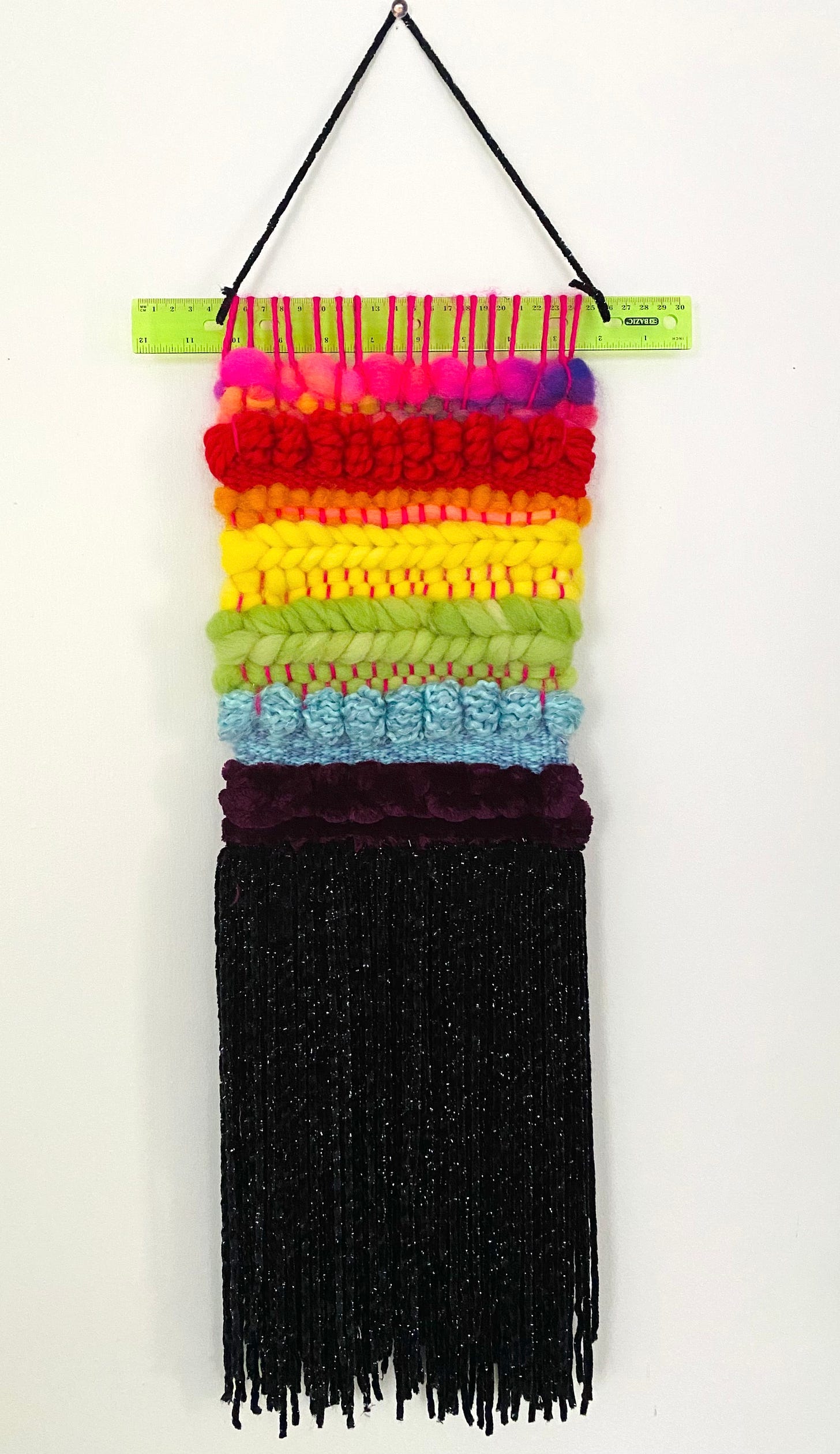 A woven wall tapestry about 2.5 feet long that is the colors of the LGBT Pride Flag from pink to black. It is hung on a lime green ruler.