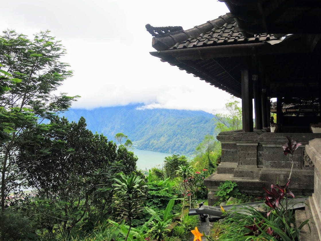 The side of a Balinese temple surrounded by lush green tropical foliage. In the distance cloud-capped mountains loom over a lake.