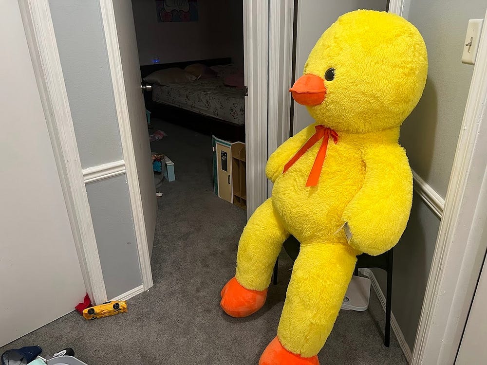 Photo of a giant duck plushie seated in a chair beside an open bedroom door.