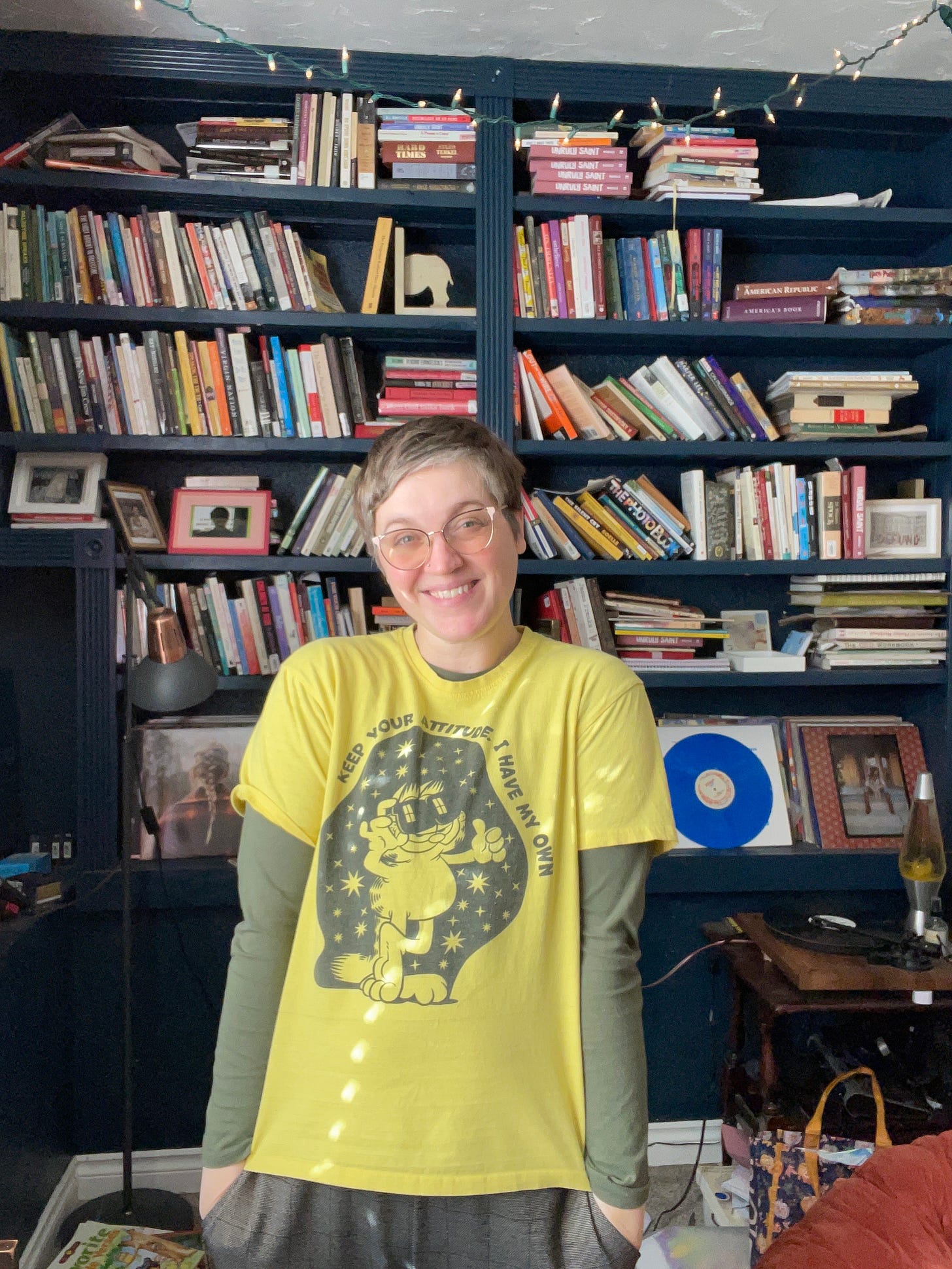 a white person with short hair and glasses stands in front of a bookshelf piled high with books and records. They are smiling and waering a yellow garfield shirt